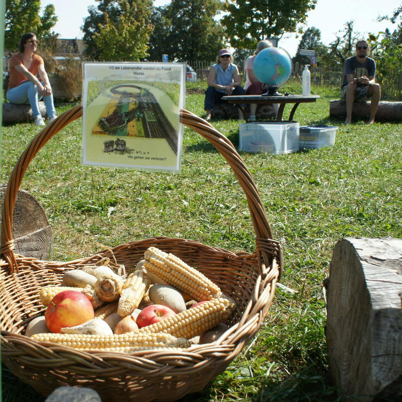 Korb mit Mais und Äpfeln, im Hintergrund sitzen Menschen in einem Kreis.
