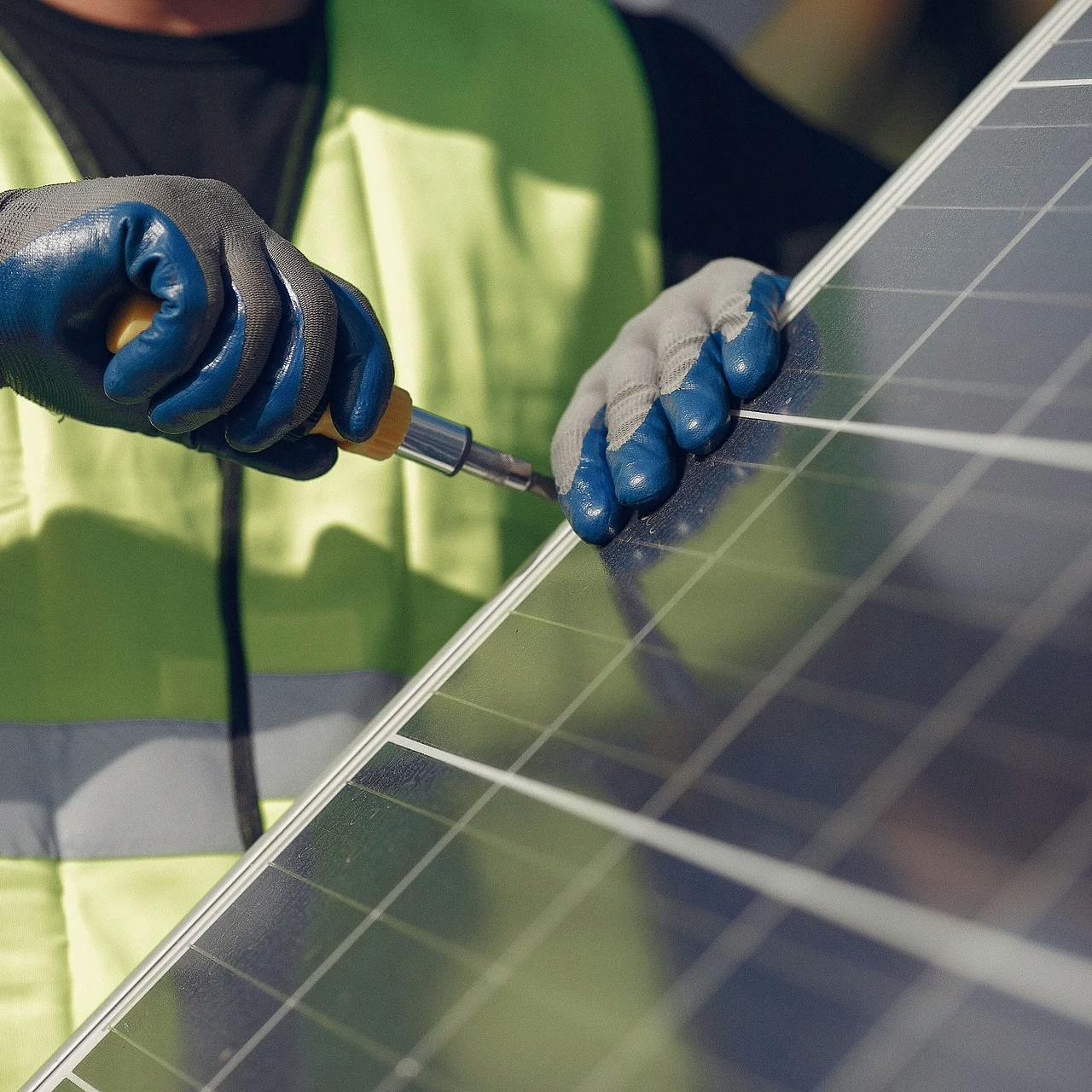 Ein Mann mit Leuchtweste in Arbeiterhandschuhen schraubt an einem Solarpanel herum.