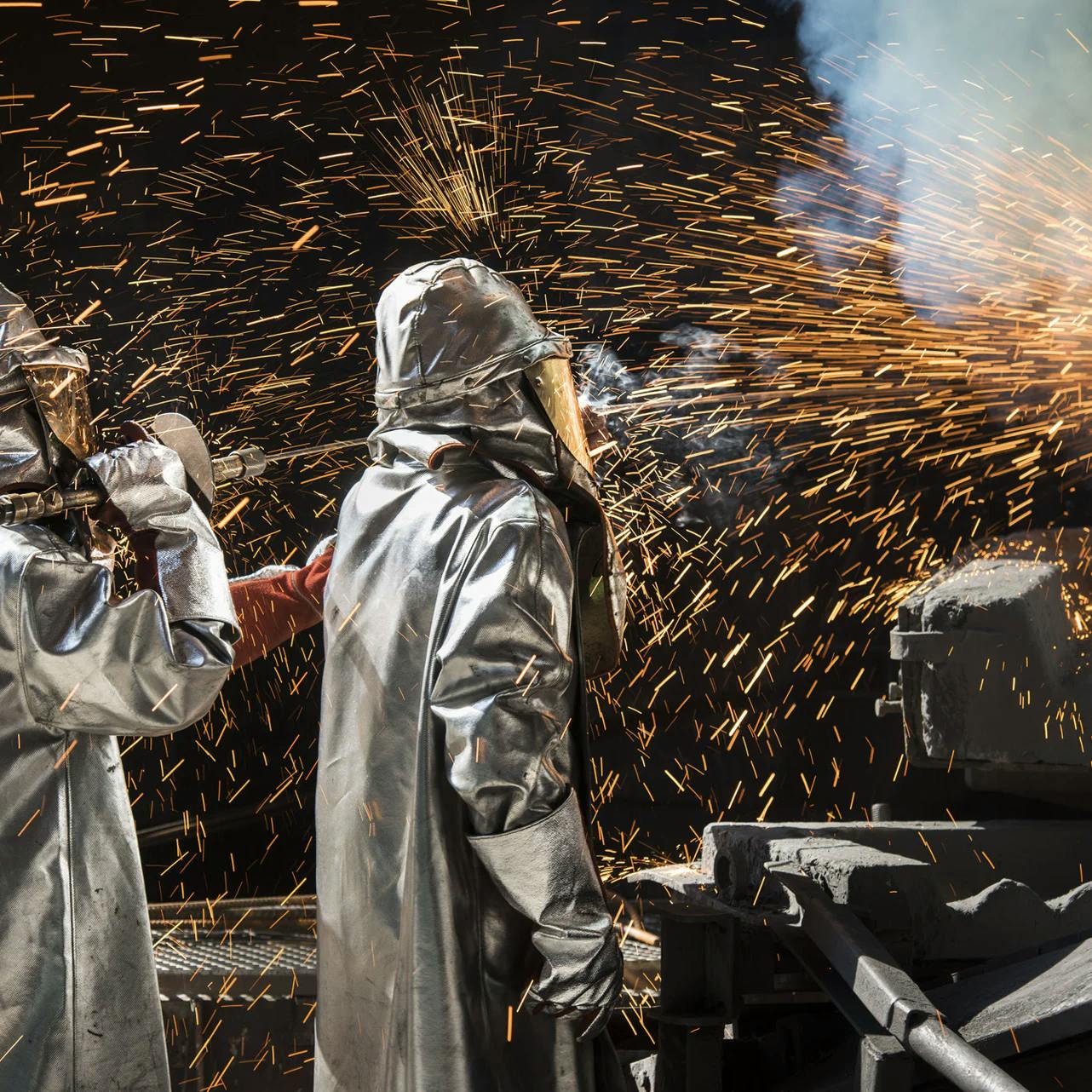 Zwei Arbeiter stehen mit Schutzkleidung vor einem Batterie-Recycling-Ofen, von dem Funken sprühen.